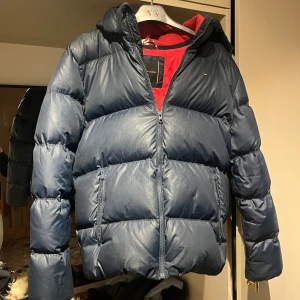 Blå pufferjacka från Tommy Hilfiger - Säljer en snygg blå pufferjacka från Tommy Hilfiger i storlek 176. Perfekt för kalla dagar med sin vadderade design. Insidan är röd vilket ger en snygg kontrast. Passar både till vardags och lite finare tillfällen! Passar både killar och tjejer, ny pris låg runt 1950. Den är 3-4 år gammal, därför säljer jag den för billigt men den är i bra skick.