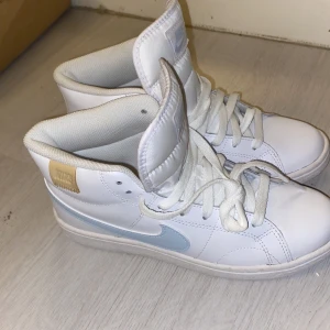 Vita Nike sneakers med ljusblå detalj - Säljer ett par snygga vita Nike sneakers i mycket bra skick. De har en ljusblå swoosh och är perfekta för både vardag och träning. Skorna har snörning och en bekväm passform. Storleken är dam 37.5 passa på att köpa dessa stilrena sneakers! Passar både män o kvinnor. Aldrig använda ute bara provat. Anledningen till säljning är kommer inte till nytta eftersom dem är förstora