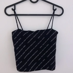 Svart axelbandslös topp med textmönster - Snygg svart axelbandslös topp med tunna axelband och ett coolt textmönster i vitt. Perfekt för en utekväll eller fest. Toppen är i bra skick och har en tight passform som framhäver figuren.