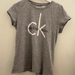 Grå t-shirt från Calvin Klein Jeans - Säljer en snygg grå t-shirt från Calvin Klein Jeans i storlek L. Den har en klassisk design med korta ärmar och det ikoniska CK-logotypen tryckt på framsidan. Perfekt för en avslappnad stil eller som basplagg i garderoben.