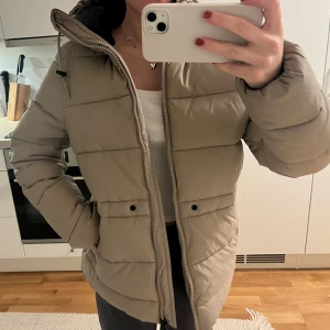 Beige pufferjacka med huva - Varm vinterjacka från Everest i mycket bra skick. Tillkommer ett snöre man har runt midjan och drar åt, vilket jag själv tagit bort. Praktisk med reflexband i fickorna som man kan ha utanför jackan om man vill.