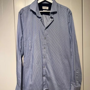 Blå skjorta från Bruun & Stengade - Säljer en stilren blå skjorta från Bruun & Stengade i slim fit. Skjortan har ett diskret randigt mönster och är tillverkad i ett lättskött material. Stryker du skjortan är den så gott som ny. Nypris 900:-