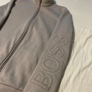 Beige/Grå ziptröja från Hugo Boss - Säljer en snygg beige/grå ziptröja från Hugo Boss i nytt skick från Hennings. Tröjan har en dragkedja framtill och BOSS-loggan broderad på ärmen. Perfekt för både vardag och lite finare tillfällen. Den är superbekväm och stilren!