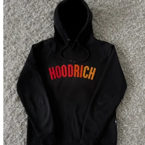 Svart hoodie från Hoodrich - Säljer en svart hoodie från Hoodrich med färgglad text på bröstet. Den har en klassisk passform med en stor ficka fram och justerbar huva. Perfekt för en avslappnad stil eller streetwear-look. Använd ett par gånger men i mycket bra skick!