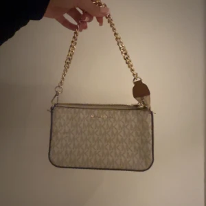 Beige axelväska med kedja från Michael kors - Snygg beige axelväska med guldfärgad kedja och dragkedja. Väskan har ett stilrent mönster och är perfekt för både vardag och fest. Den är i mycket bra skick och har en praktisk storlek för att bära med sig det viktigaste.