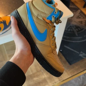 Nike COURT BOROUGH MID 2 BOOT GS - Säljer ett par snygga beige Nike sneakers med blå detaljer. Skorna har en klassisk design med snörning och en bekväm passform. Perfekta för både vardag och träning. De är i mycket bra skick och redo för nya äventyr!