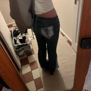 Blå jeans med slitna detaljer - Säljer ett par snygga blå jeans med slitna detaljer på bakfickorna. De är lågmidjade och har en bootcut-stil som ger en retro vibe.