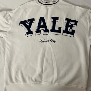 Vit sweatshirt med Yale-tryck - Säljer en vit sweatshirt med stort Yale University-tryck på framsidan och siffran 19 på ärmen. Perfekt för en avslappnad stil eller för att visa ditt stöd för Yale! Tröjan har en normal passform och är gjord i mjukt bomullsmaterial.
