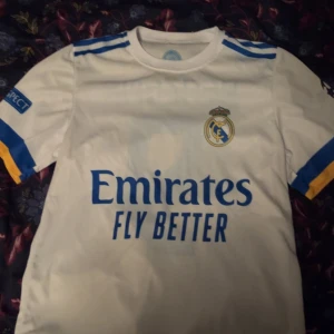 Vit Real Madrid tröja och blå PSG tröja - Säljer två fotbollströjor i bra skick! En vit Real Madrid tröja med korta ärmar och blå detaljer, samt Benzema #9 på ryggen. Den andra är en blå PSG tröja med korta ärmar, röd och vit rand, och Neymar Jr #10 på ryggen. Perfekt för fotbollsfans! ⚽300kr för båda tröjorna!