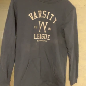 Mörkblå hoodie med tryck - Säljer en mörkgrå hoodie med texten 'Varsity League 1996' tryckt på framsidan. Den har en klassisk känguruficka och långa ärmar. Knappt använd.
