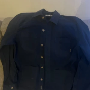 Mörkblå overshirt - Säljer en stilren mörkblå skjorta med knappar. Skjortan har långa ärmar och två bröstfickor. Perfekt för både vardag och festliga tillfällen. Materialet känns slitstarkt och bekvämt. Passar S och M använd bara 4 gånger Max .