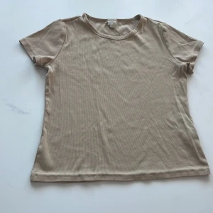 Beige ribbad t-shirt - Säljer en snygg beige ribbad t-shirt i mycket bra skick. Den är kortärmad och perfekt för en avslappnad stil. Materialet är mjukt och stretchigt, vilket gör den superbekväm att ha på sig. Passar perfekt till både jeans och kjol. Perfekt för vår och sommar!
