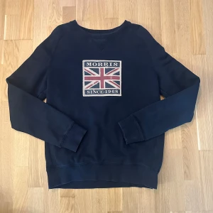Morris sweatshirt - Mörkblå tröja med en stor, tryckt flagga på framsidan. Tröjan har en rund halsringning och långa ärmar. Tröjan är i fint skick men lite blekt i kragen, se bilder. Sedan lite smått sliten i några sömmar (se bild 3 och 4). Storlek M. Herr