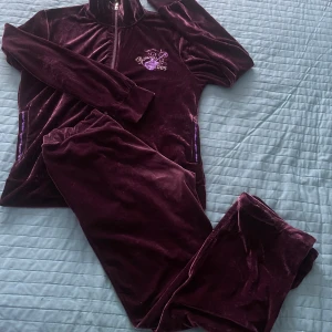 Mörklila velourset - Säljer ett snyggt och bekvämt mörklila velourset med dragkedja och broderad detalj på tröjan.  Tröjan har långa ärmar och byxorna har något kortare passform. 