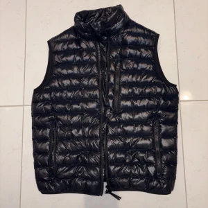 Svart dunväst från Peak Performance - Säljer en snygg svart dunväst från Peak Performance i mycket bra skick. Västen har en quiltad design med dragkedja framtill och två praktiska fickor. Perfekt för höst och vår när du vill hålla dig varm utan att bära en jacka. Passar både till vardags och lite mer uppklädda tillfällen. (Finns ej i sortiment längre)
