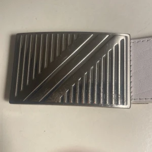 Vitt skärp från Saddler - Snyggt vitt skärp i äkta läder från Saddler med ett silverfärgat spänne med mönster. Skärpet har flera hål för justerbar passform. 