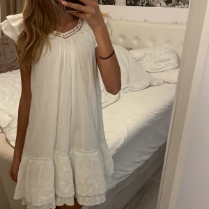 Vit klänning från Next - Säljer en superfin klänning från Next. Den har en söt volangdetalj längst ner och vackra broderier. Halsringningen är dekorerad med flätade detaljer och små pärlor. Perfekt till midsommar!Klänningen är ärmlös och har en lös passform. Inkluderar ett vitt underlinne.