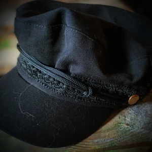 Svart keps med dekorativt band - Snygg svart keps med ett dekorativt band och en liten guldfärgad knappdetalj på sidan. Perfekt för att ge din outfit en stilfull touch. Kepsen har en klassisk design och passar till många olika tillfällen.