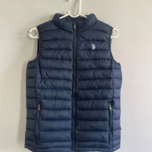 Mörkblå dunväst med dragkedja - Säljer en snygg mörkblå Polo dunväst  Perfekt för höst och vår, den är både stilren och praktisk. Västen har en normal passform och är i nyskick. Västen har en extremt liten skavank som inte riktigt syns med blotta ögat men kan skicka bild om ni är intresserade!