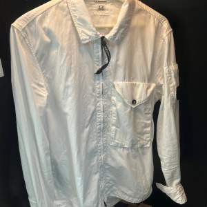 Säljer en stilren vit Overshirt från C.P. Company. Overshirten har en cool detalj med en lins på ärmen och en bröstficka med knapp. Perfekt för både vardag och fest. Den är i bra skick och passar till många olika outfits!