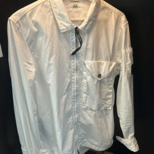 Vit långärmad OVERSHIRT från C.P. Company - Säljer en stilren vit Overshirt från C.P. Company. Overshirten har en cool detalj med en lins på ärmen och en bröstficka med knapp. Perfekt för både vardag och fest. Den är i bra skick och passar till många olika outfits!