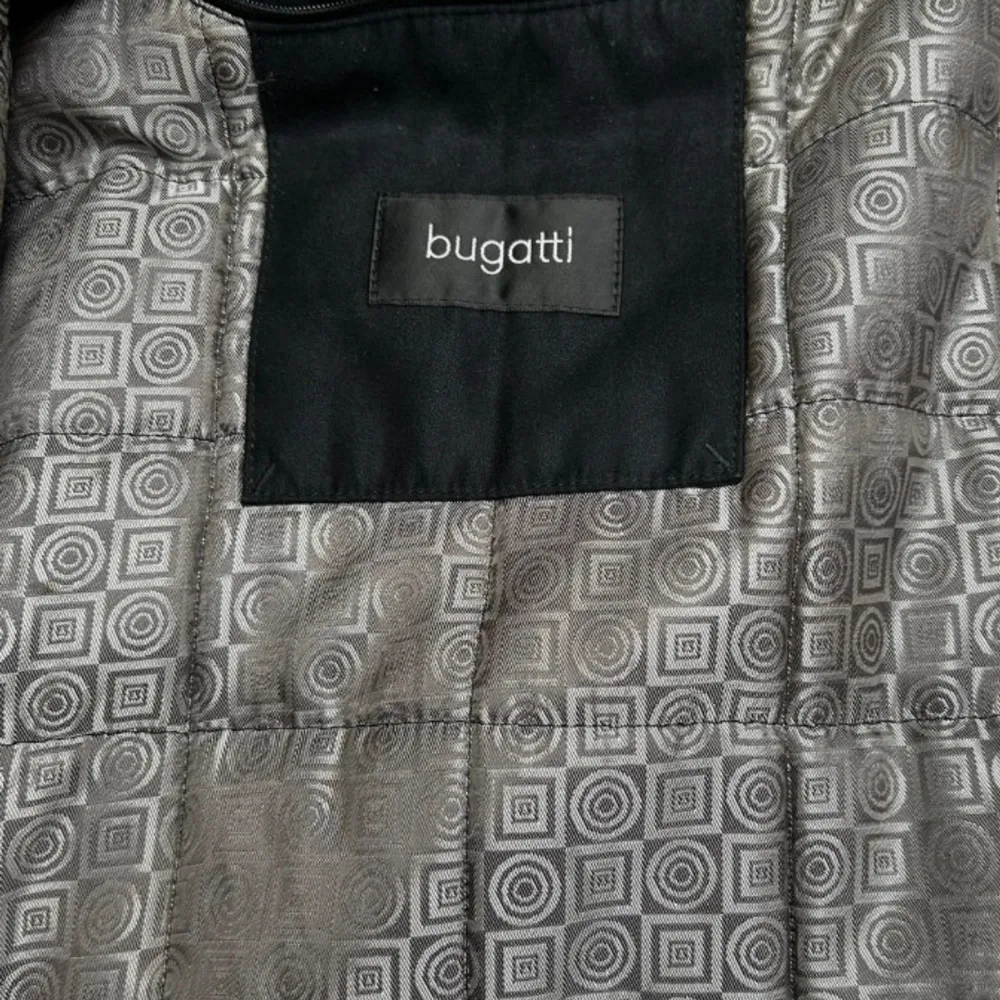 Säljer en stilren svart jacka från Bugatti i storlek 27. Jackan har långa ärmar och stängs med knappar. Den är tillverkad i 100% polyester och har ett snyggt mönstrat foder. Perfekt för höst och vinter!. Jackor.