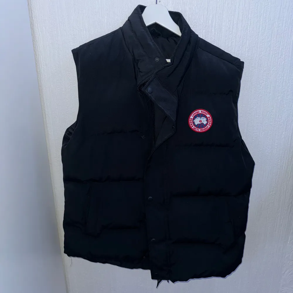 Säljer en svart dunväst från Canada Goose i storlek L. Västen har en klassisk design med knappar och dragkedja framtill. Perfekt för kyliga höst- och vinterdagar. Den är i nyskick och har en snygg logga på bröstet. Passar både till vardags och lite mer uppklädda tillfällen.. Jackor.