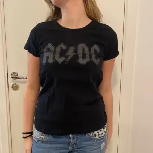 Säljer en svart AC/DC t-shirt i bomull. Den har ett coolt tryck med bandets logga i grått på framsidan. Perfekt för rockälskare och passar till både vardag och konsert. T-shirten är kortärmad och har en normal passform.