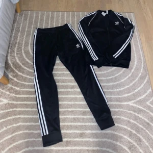 Svart träningsset från Adidas - Säljer ett svart träningsset från Adidas med de klassiska tre vita ränderna längs ärmar och ben. Setet består av en jacka med dragkedja och matchande byxor. Perfekt för träning eller en avslappnad dag. Superbekvämt och stilrent! 🖤