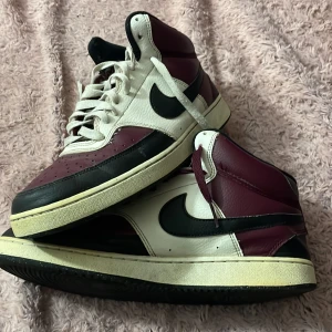 Nike sneakers i vinrött och vitt - Säljer ett par coola Nike sneakers i vinrött, vitt och svart. De har en klassisk design med snörning och är perfekta för både vardag och fest. Skorna är i skinn och har en retro vibe som är supertrendigt just nu. Passar perfekt till jeans eller en avslappnad outfit.