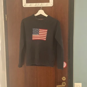 Gant stickad tröja - Säljer en snygg mörkblå stickad tröja med en stor amerikansk flagga på framsidan. Tröjan har långa ärmar och ribbade muddar vid ärmslut och nederkant. Perfekt för höst och vinter!