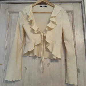 Beige kofta från Gina Tricot - Säljer en söt beige kofta från Gina Tricot med volangdetaljer och knytning framtill. Koftan har långa ärmar och är perfekt för en chic look. Passar bra till både jeans och kjol. Storlek XS