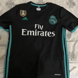 Svart Real Madrid fotbollströja från Adidas - Säljer en svart Real Madrid fotbollströja från Adidas med korta ärmar. Tröjan har turkosa detaljer och klubbens emblem på bröstet. Den har även Fly Emirates som sponsorlogga och ett märke för FIFA Club World Cup. Perfekt för fans av laget! Pris kan diskuteras!!