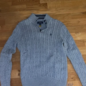 14-18 år Grå stickad tröja från Polo Ralph Lauren storlek 14-18 år - Snygg grå stickad tröja från Polo Ralph Lauren med dragkedja vid halsen. Tröjan har ett diskret broderat märke på bröstet och är perfekt för kyligare dagar. Den har långa ärmar och en klassisk passform.