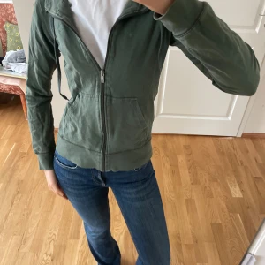 Grön zip-up hoodie - Säljer en grön zip-up hoodie med dragkedja och fickor framtill. Den har långa ärmar och en bekväm passform, perfekt för en avslappnad stil. Hoodien är tillverkad i ett mjukt material som känns skönt mot huden.