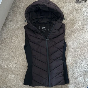 Svart dunväst från Zara - Snygg svart dunväst från Zara med huva och dragkedja framtill. Perfekt för lager-på-lager under kyliga dagar. Västen har en stilren design med quiltade detaljer.