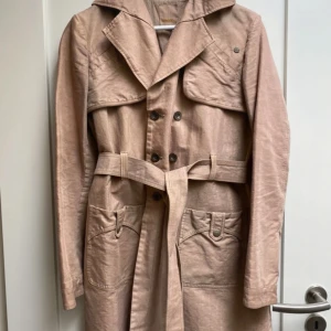 Beige trenchcoat från Diesel - Snygg beige trenchcoat från Diesel med knappar och bälte i midjan. Jackan har långa ärmar och praktiska fickor framtill. Perfekt för en stilren look under höst och vår.