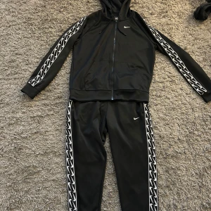 Svart träningsset från Nike - Säljer ett svart träningsset från Nike med dragkedja och huva. Setet har vita mönster längs ärmar och ben samt små Nike-loggor. Perfekt för träning eller en avslappnad dag.