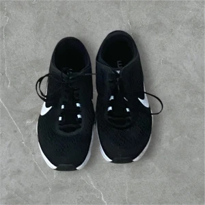 Svarta Nike sneakers - Snygga svarta Nike sneakers med vit sula och klassisk swoosh-logga på sidan. Skorna har snörning och en luftig mesh-design som ger bra ventilation. Perfekta för både träning och vardag.