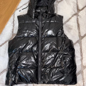 Svart glansig dunväst med huva - Snygg svart glansig dunväst med dragkedja och huva. Perfekt för kyligare dagar när du vill hålla stilen. Västen har praktiska fickor framtill och en quiltad design som ger extra värme.