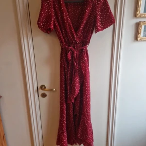 Röd prickig klänning med knytband - Charmig röd klänning med vita prickar och v-ringning. Den har korta ärmar och ett knytband i midjan för en smickrande passform. Perfekt för en somrig look!