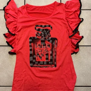 Röd topp med volangärmar och parfymtryck storlek M - Snygg röd topp med volangärmar och ett coolt parfymtryck i svart och vitt på framsidan. Perfekt för att ge en färgklick till din outfit!