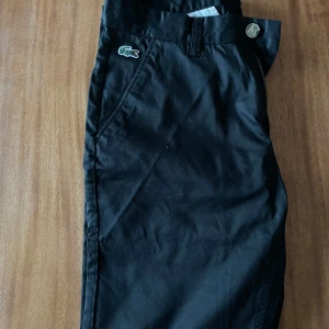 Lacoste chinos  - Snygga svarta byxor från Lacoste i slim fit. Byxorna har en klassisk design med dragkedja och knapp framtill. Perfekta för en stilren look.