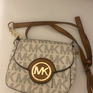 Beige axelväska från Michael Kors - Snygg beige axelväska från Michael Kors med det ikoniska MK-mönstret. Väskan har en justerbar axelrem i brunt läder och guldfärgade detaljer. Perfekt för att ge din outfit en lyxig touch.