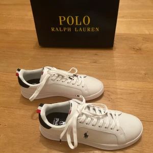 Vita sneakers från Polo Ralph Lauren - Aldrig använda med lappar kvar! As snygga vita sneakers från Polo Ralph Lauren med klassisk design. Skorna har snörning och en diskret logga på sidan. De har en vit sula och detaljer i rött, guld och blått på hälen i mocka.  Perfekta för en stilren look. Har fysiskt och digitalt kvitto. Nypris 1595:-. Pris kan diskuteras vid snabb affär! Passa på nu när det är gratis frakt