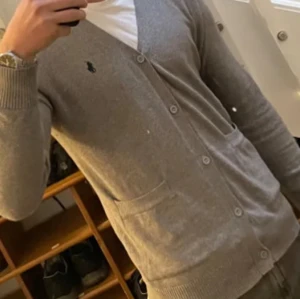 Grå kofta från Ralph Lauren - Säljer en stilren grå kofta från Ralph Lauren med knappar framtill och en broderad logotyp på bröstet. Koftan har långa ärmar och ribbade muddar vid ärmslut och nederkant. Perfekt för en klassisk look.