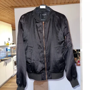 Svart bomberjacka från The Black Label Collection - Snygg svart bomberjacka från The Black Label Collection i storlek S. Jackan har en dragkedja framtill och en praktisk ficka med dragkedja på ärmen. Perfekt för en stilren look.