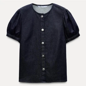 Mörkblå jeansblus från Zara - Snygg mörkblå jeansblus från Zara med korta puffärmar och knappar framtill. Perfekt för en stilren look med en touch av denim. Blusen har en rund halsringning och är gjord i ett bekvämt material.