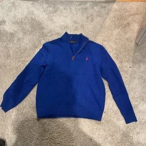 Blå tröja med dragkedja från Ralph Lauren - Snygg blå tröja från Ralph Lauren med dragkedja vid halsen och det klassiska logotypbroderiet på bröstet. Tröjan har långa ärmar och ribbade muddar vid ärmslut och nederkant. Perfekt för en stilren look.