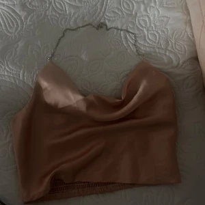 Beige axelbandslös topp från Shein - Säljer en stilren beige axelbandslös topp från Shein. Toppen har en elegant kedja som axelband och en smickrande drapering framtill. Perfekt för en kväll ute eller en speciell tillställning.